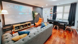 Una sala moderna con un sofá gris, cojines coloridos, una silla de mimbre, una mesa de comedor y una pantalla grande mostrando la interfaz de Netflix.