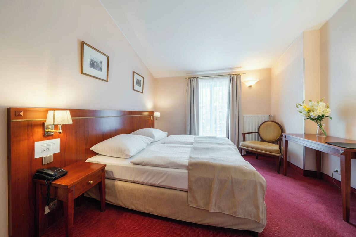 A hotel room with a large bed, bedside table with a lamp, a chair, a desk, and a vase of flowers.