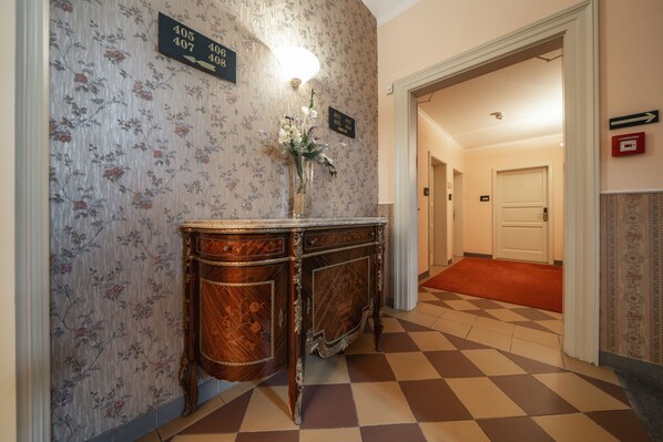 A hallway with a checkered floor, a floral wallpaper, a wooden cabinet with a vase of flowers, and directional signs. - Hotel Raffaello Prague (Prague)