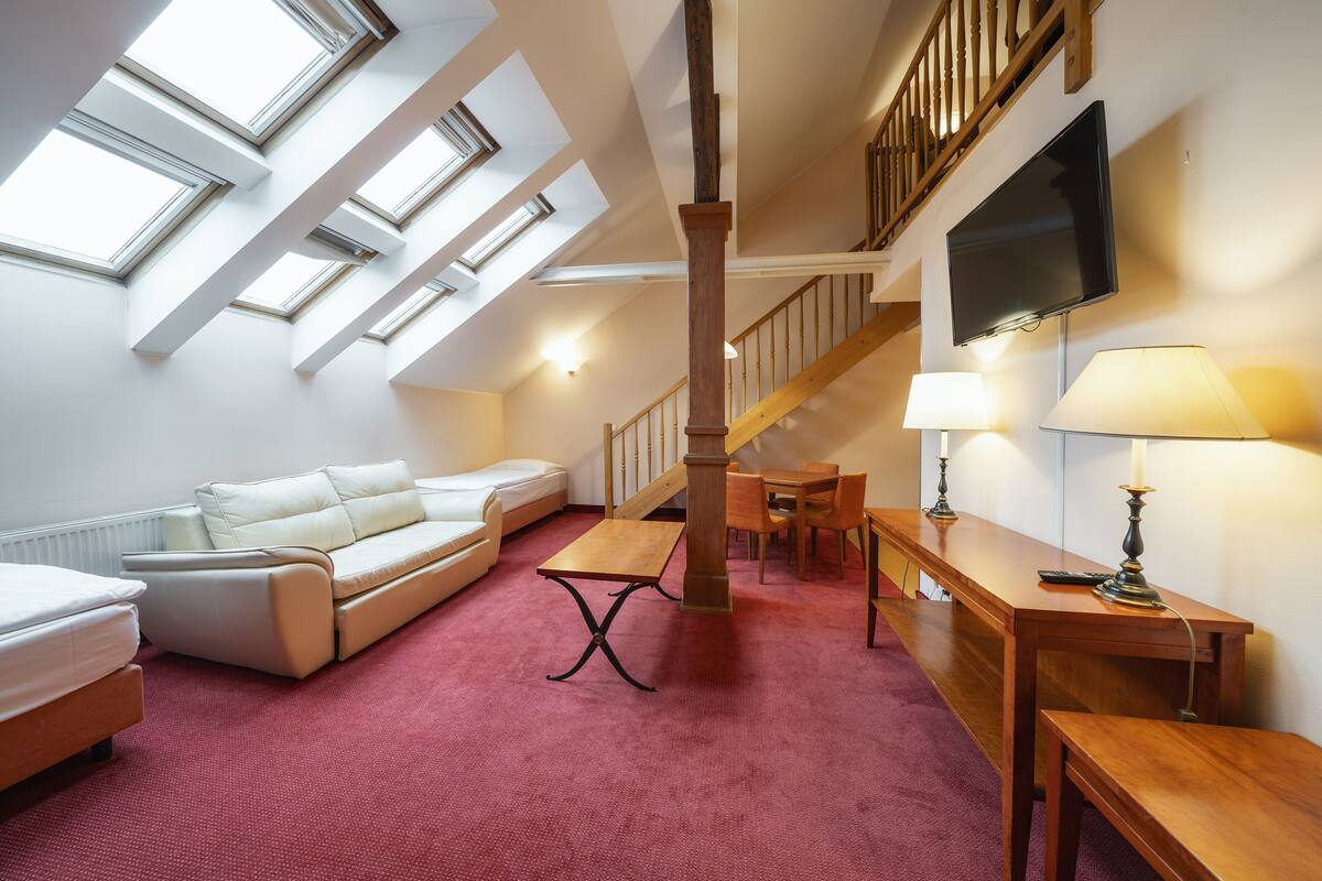 A spacious loft bedroom with skylights, a wooden staircase, a flat-screen TV, and a cozy seating area with a sofa and a small table.
