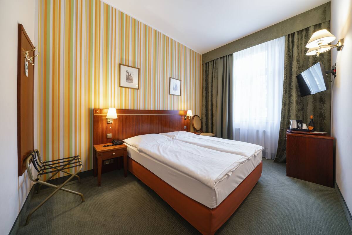 A hotel room with a large bed, two bedside lamps, a wooden nightstand, a chair, a desk, a television, and striped wallpaper.