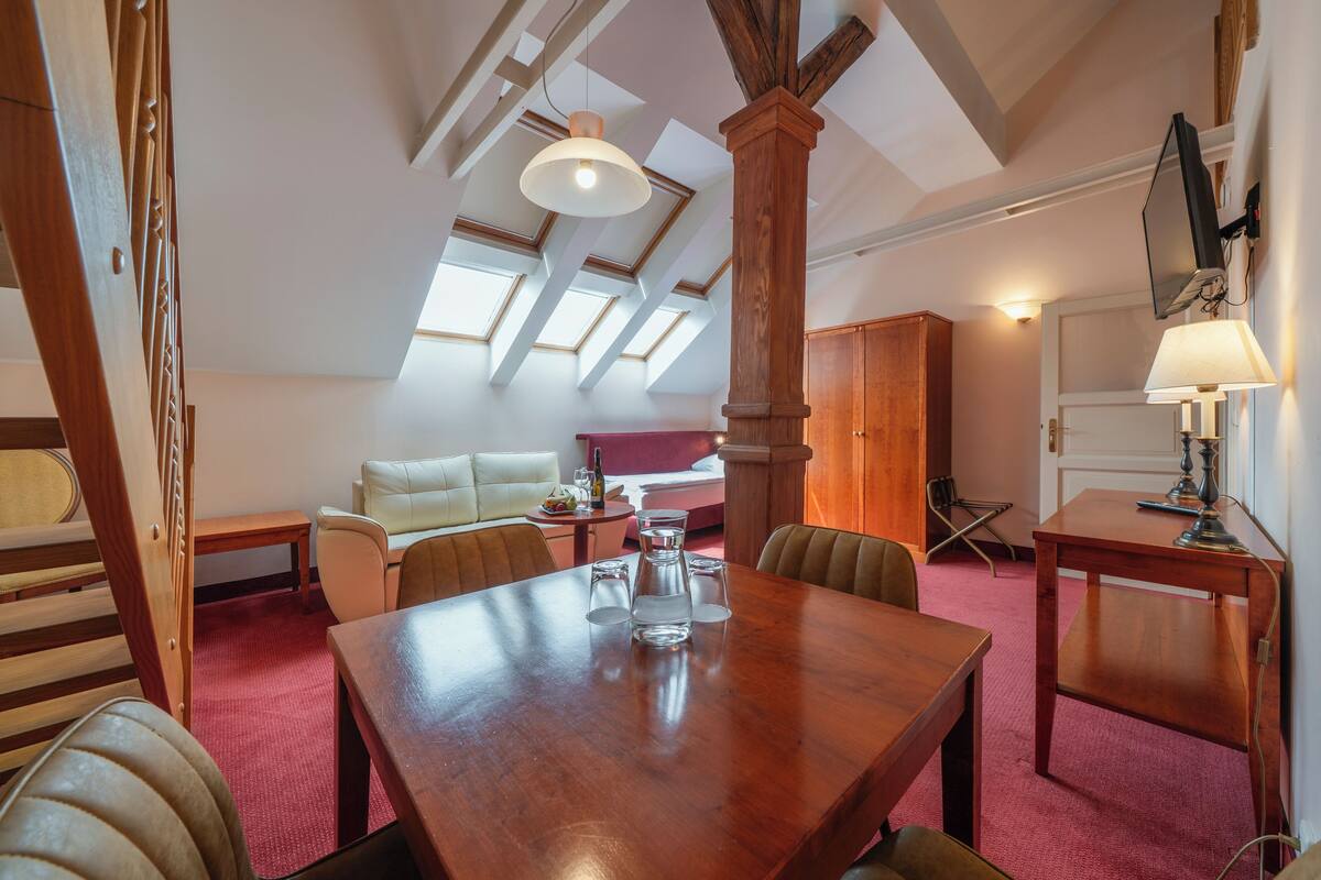 A dining area with a wooden table, chairs, and a red carpet. There is a wooden bench and a small table with a lamp. The room has a sloped ceiling with skylights and a wooden column.