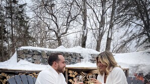 Dos personas sentadas junto a una fogata con leña, en un entorno nevado al aire libre.