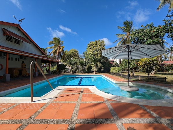 A swimming pool with a tiled deck, a covered area with a striped umbrella, and a building with a red roof and white walls. - Capi´s Place (San Andrés)