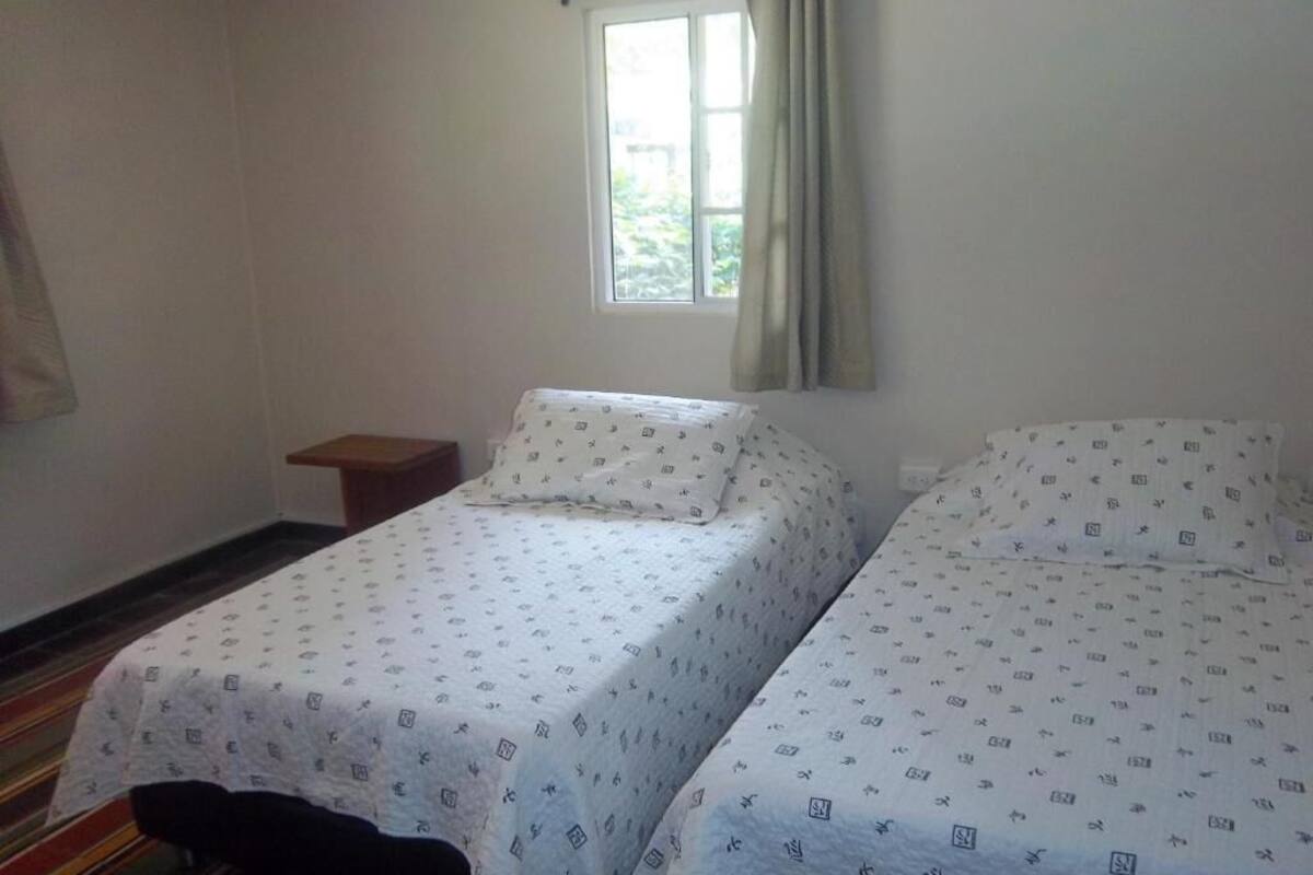 A room with two beds, a window with curtains, a small wooden nightstand, and a striped carpet.