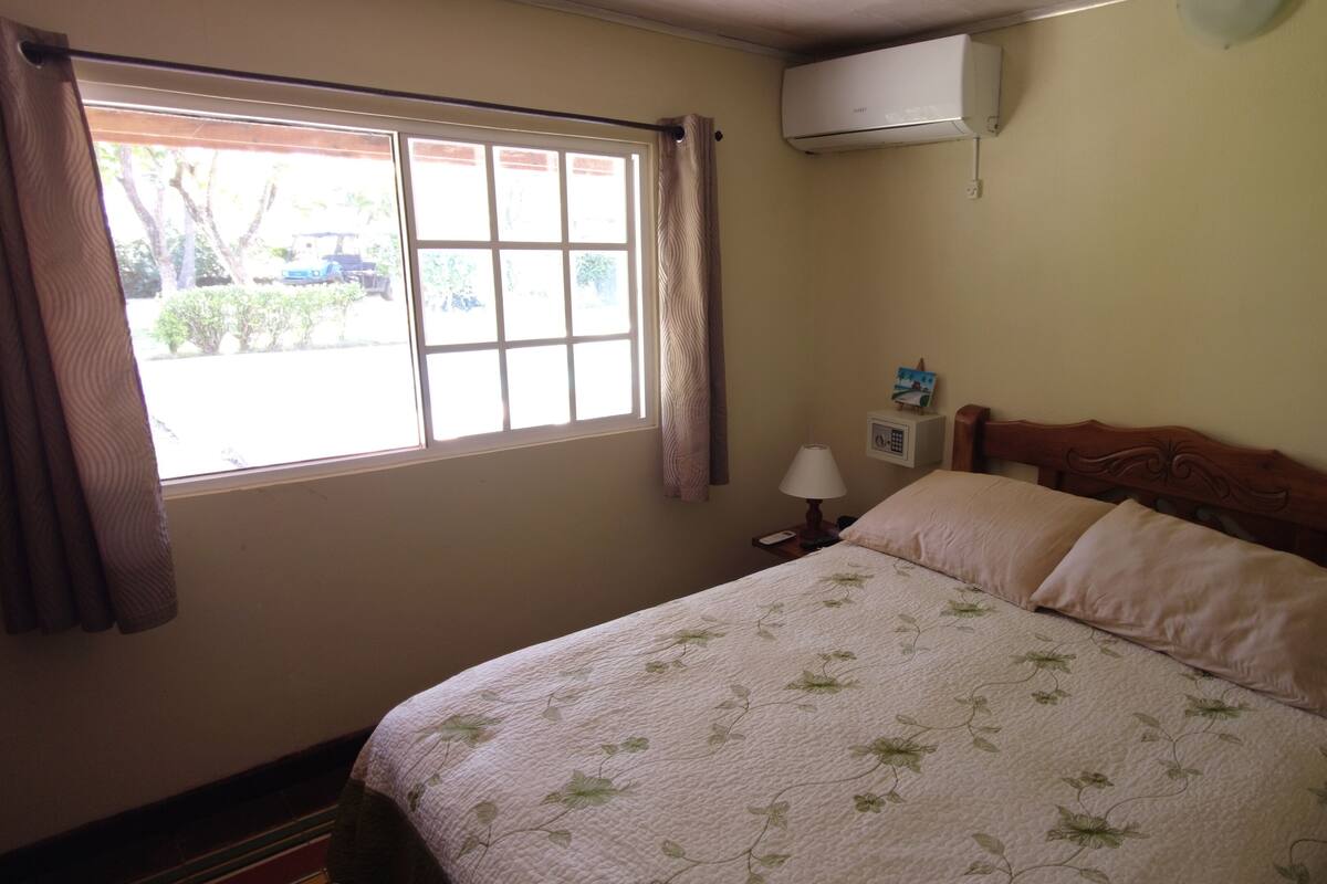 A bedroom with a bed, a nightstand, a lamp, a window with curtains, and an air conditioning unit.