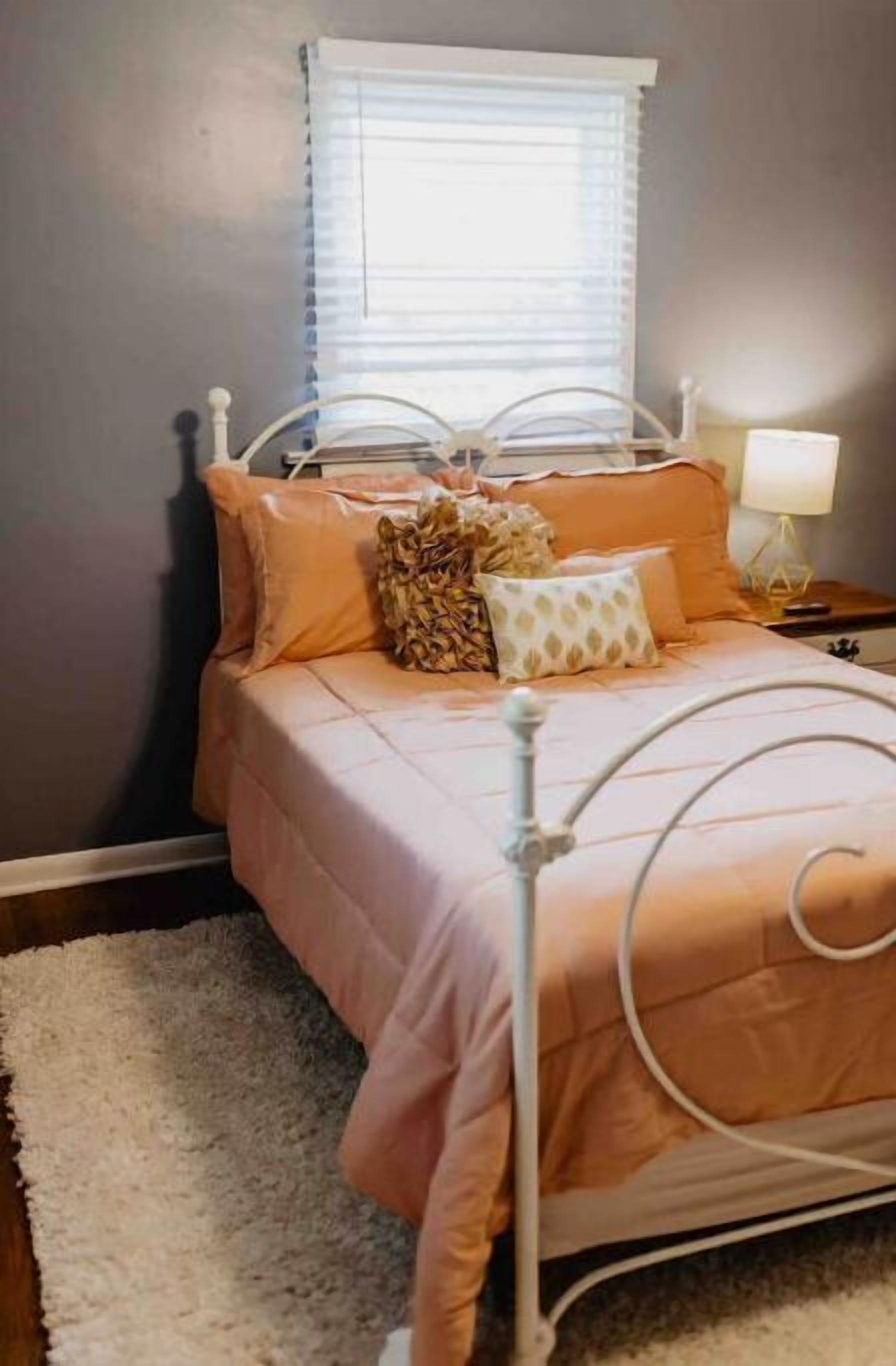 Un dormitorio con cama, almohadas, una mesita de noche y una ventana con persianas.