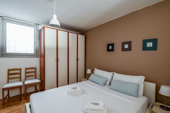 A bedroom with a bed, two chairs, a wardrobe, and a window.