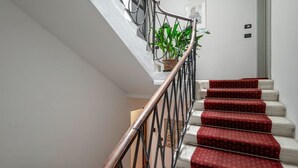 Un escalier avec une rampe en métal, des marches recouvertes d’une moquette rouge et un pot de fleurs au sommet.