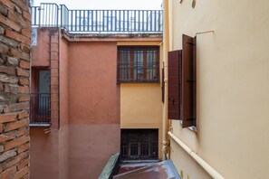 Un callejón estrecho con un muro de ladrillo a un lado y un muro amarillo al otro. Se ve una ventana con barrotes y una puerta con manija.