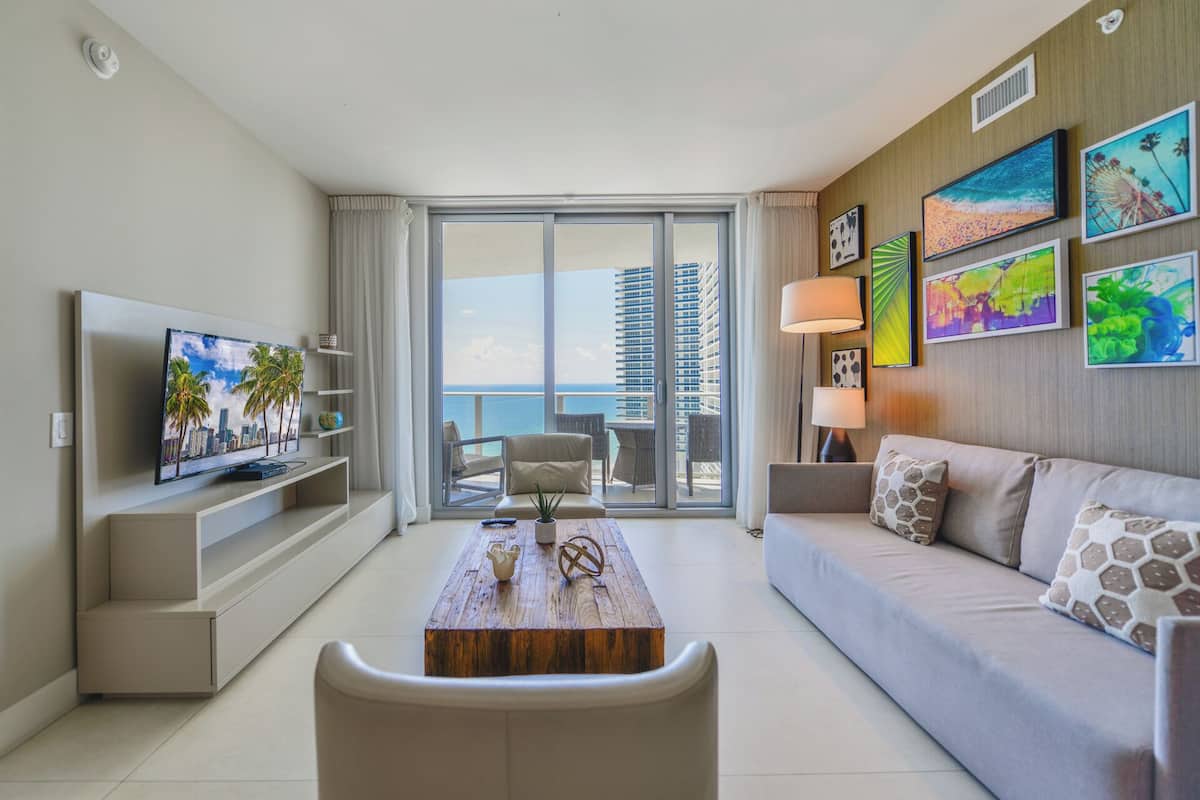A modern hotel room with a balcony, a flat-screen TV, a sofa, a coffee table, and artwork on the walls.