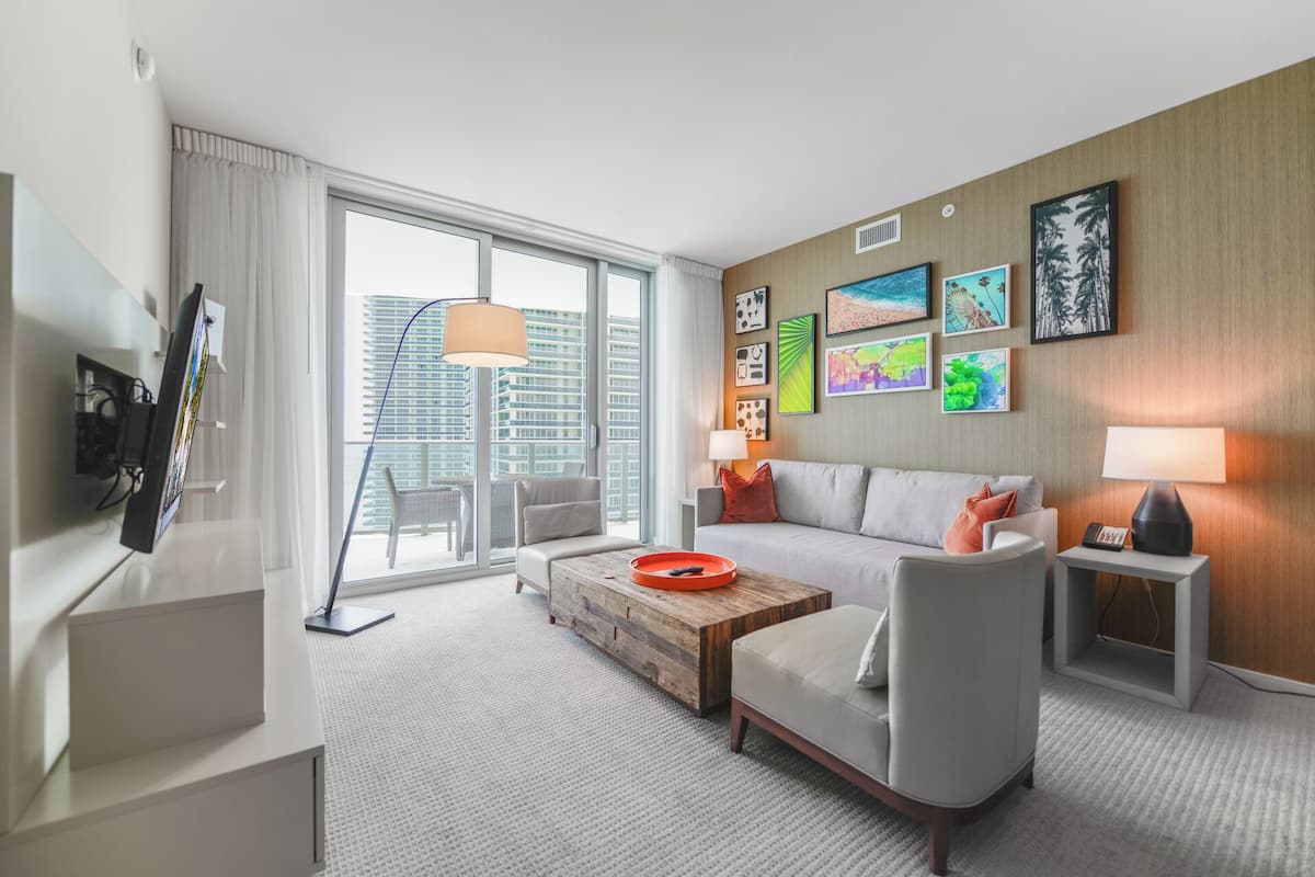 A modern hotel room with a large window, a flat-screen TV, a sofa, a coffee table, and a small side table with a lamp.