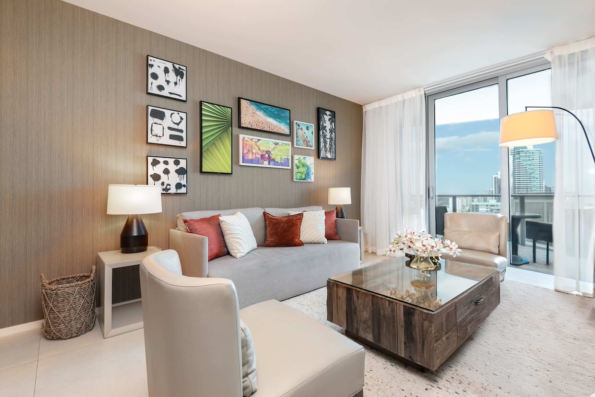 A modern hotel room with a grey sofa, a glass coffee table, and a city view through large windows.