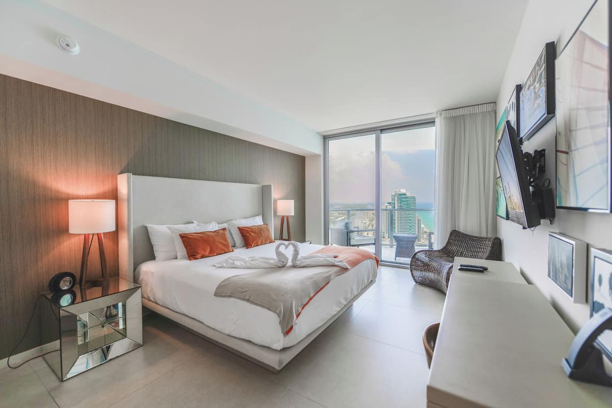 A modern hotel room with a large bed, a desk, a chair, and a city view.