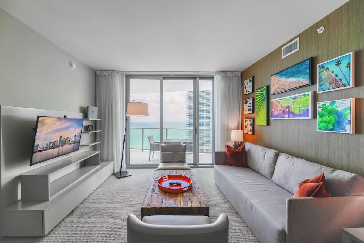 A modern hotel room with a balcony, a flat-screen TV, a sofa, a coffee table, and artwork on the walls.