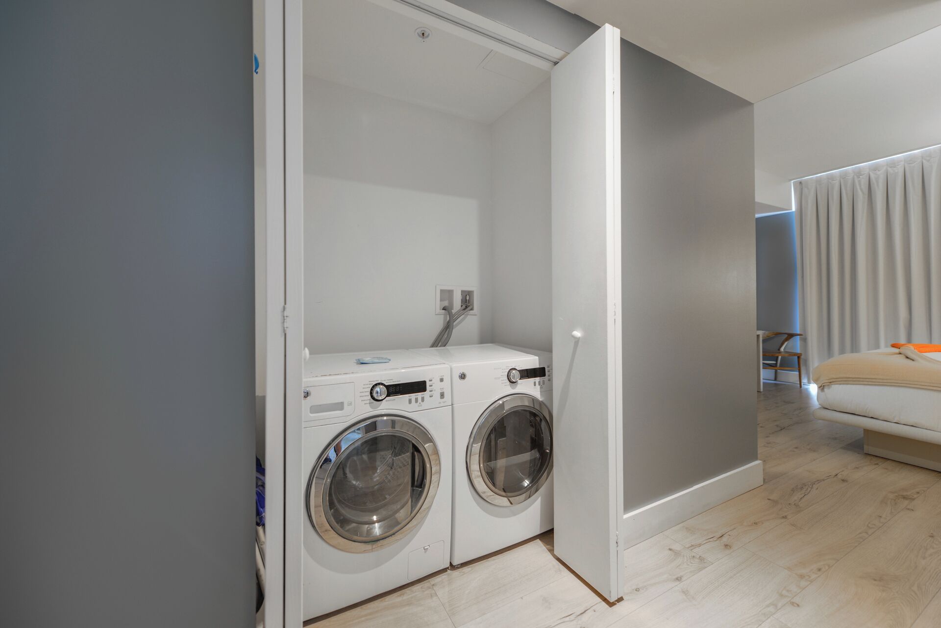 A compact laundry room with two front-loading washing machines and a dryer, adjacent to a bedroom with a bed and a window.