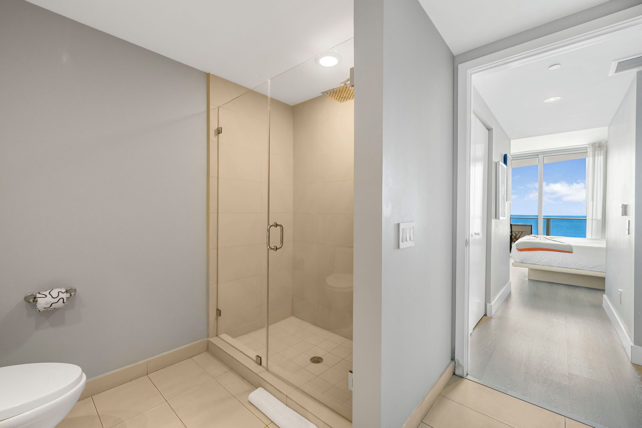 Un bagno moderno con cabina doccia in vetro, un water e una finestra che dà su una camera da letto con un letto e vista sul cielo.