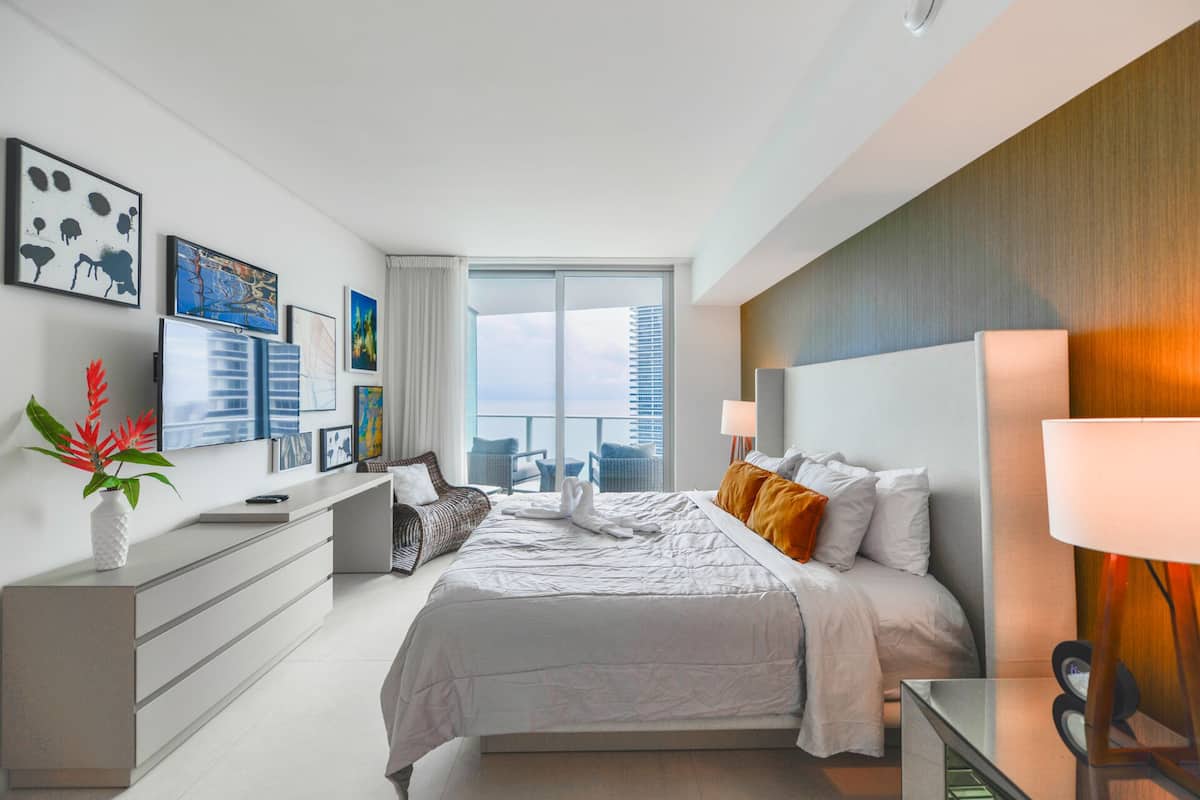 A modern hotel room with a large bed, a desk, a chair, and a view of the city.