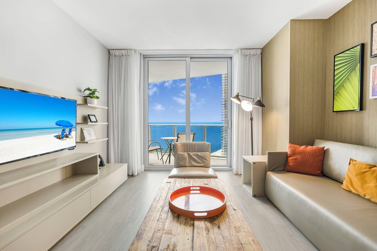 A modern living room with a large TV, a sofa, a coffee table, and a balcony with a view of the ocean.