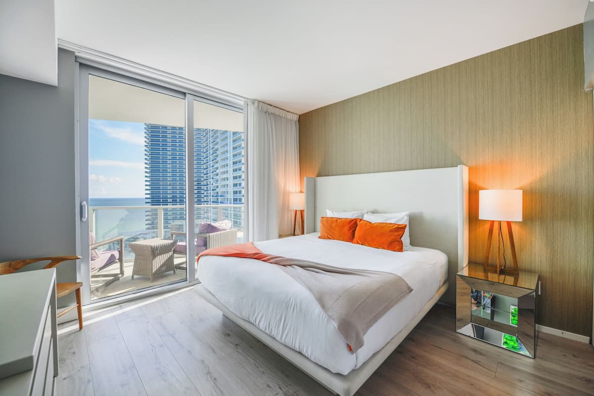 A hotel room with a large bed, a view of tall buildings, a balcony with wicker furniture, and a modern lamp.