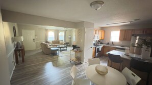 Un vaste salon comprenant un canapé, une table basse et un coin repas. On peut apercevoir une cuisine avec des meubles en bois et un réfrigérateur.