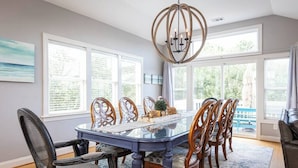 A dining area with a glass table surrounded by wooden chairs, a chandelier, and large windows with a view. - Duck Location! Volleyball, Pool, Game Room & More (Duck)