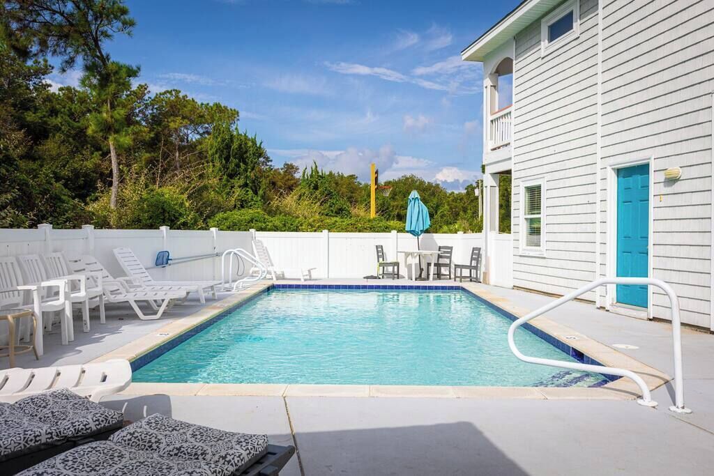 Une piscine rectangulaire dont le bord est recouvert de carreaux bleus, entourée de chaises longues blanches et d’une table avec des chaises.