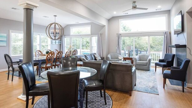 A spacious living room with a round dining table, leather chairs, a fireplace, and large windows offering a view of the outdoors. - Duck Location! Volleyball, Pool, Game Room & More (Duck)