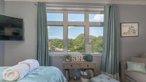A room with a bed, a desk with a lamp, a window with curtains, and a wall-mounted TV. - Cozy Resort - Pickleball, Beaches, Boat Ramps (Huddleston)