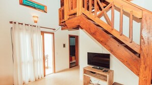 A spacious living room with a wooden staircase, a television on a wooden stand, and a door leading to another room. - Ibirawave (Imbituba)
