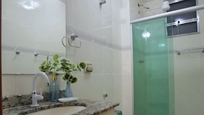 A modern bathroom with a granite countertop, a toilet with a blue seat, a glass shower enclosure, and a small window above the shower. - Mar Doce Lar (Cabo Frio)