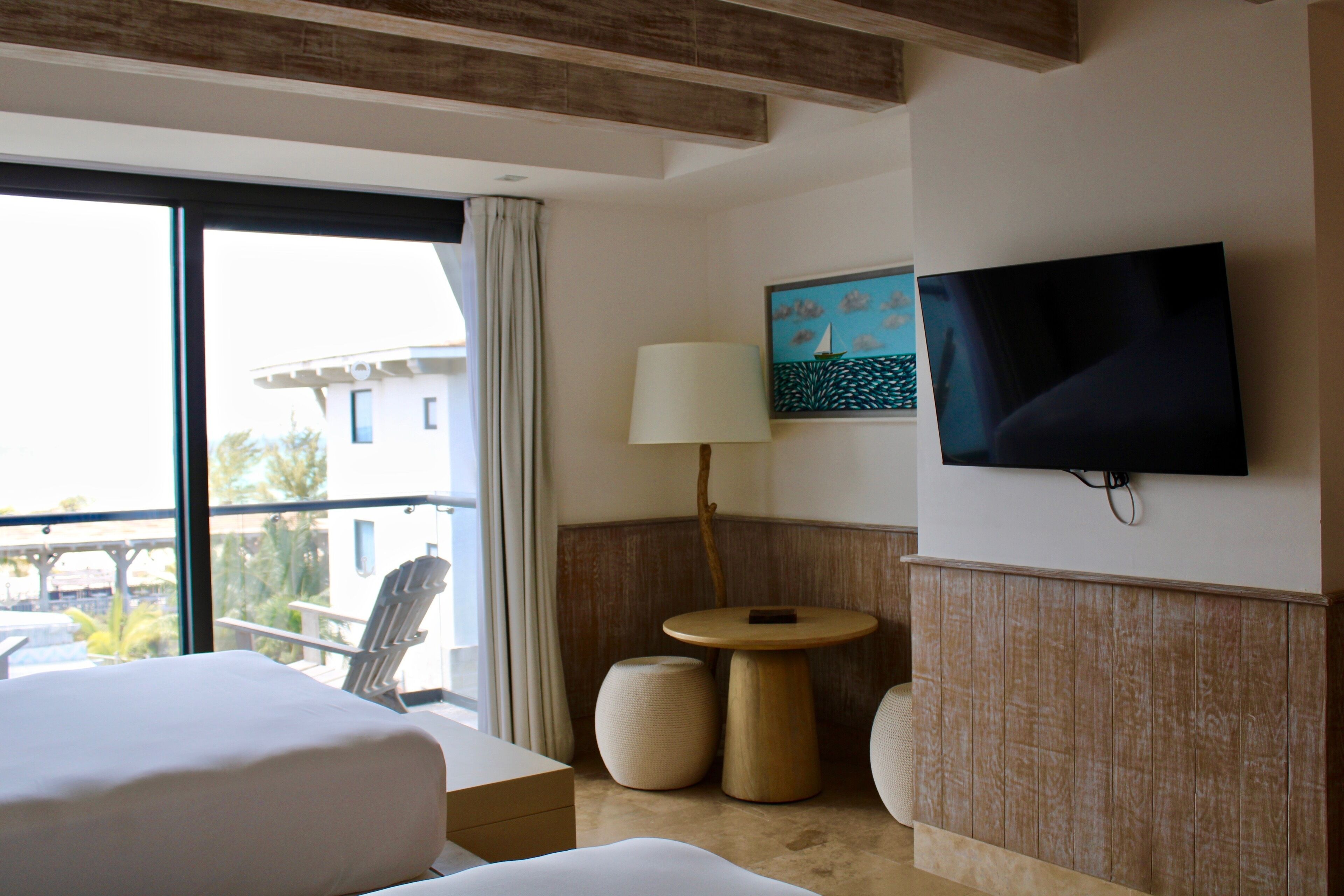 A hotel room with a bed, a round table with a lamp, a TV mounted on the wall, and a view of the outside.