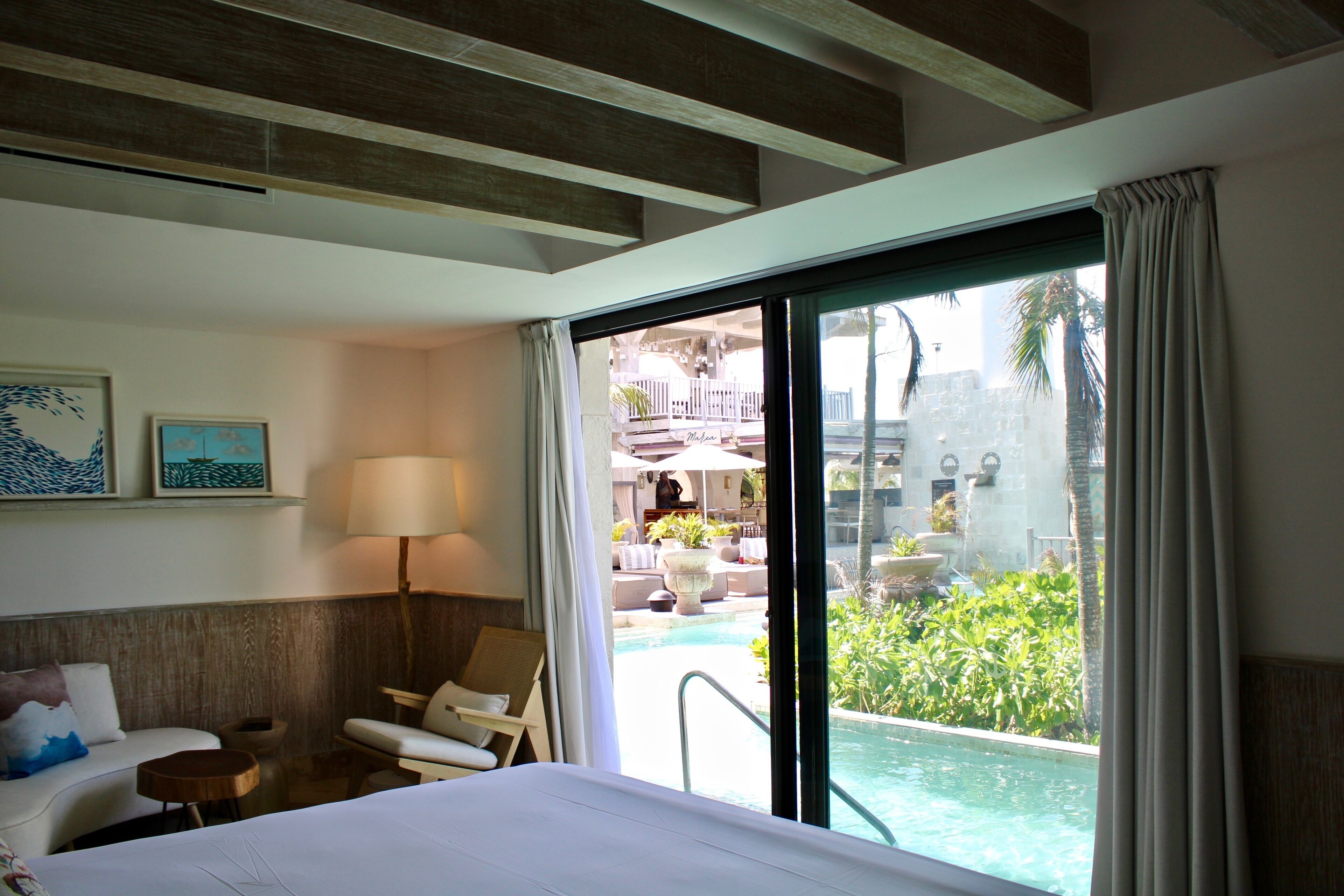 A hotel room with a bed, a desk, a chair, a lamp, and a view of a pool and outdoor area.