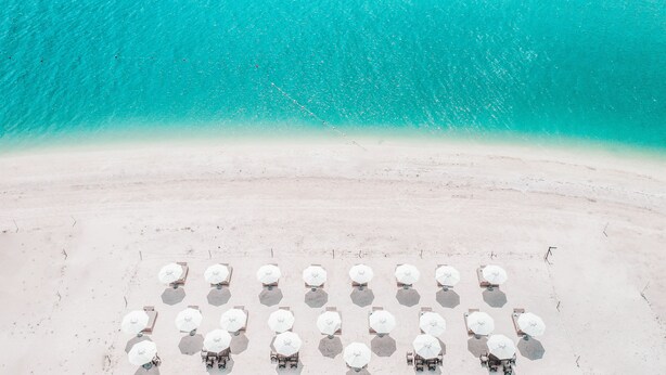 Vista aérea de una playa con hileras de tumbonas hexagonales y sombrillas, un mar turquesa cristalino y una orilla arenosa.