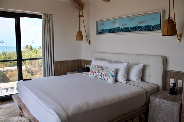 A neatly made bed with a patterned pillow, a painting of a sailboat on the wall, and a view of the ocean through a large window. - Delek Holbox (Isla Holbox)
