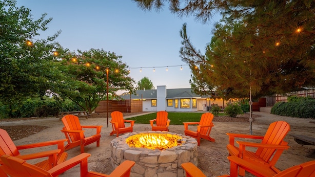 Ein Lagerfeuer, umringt von orangefarbenen Adirondack-Stühlen, in einem Garten mit einem Haus im Hintergrund.