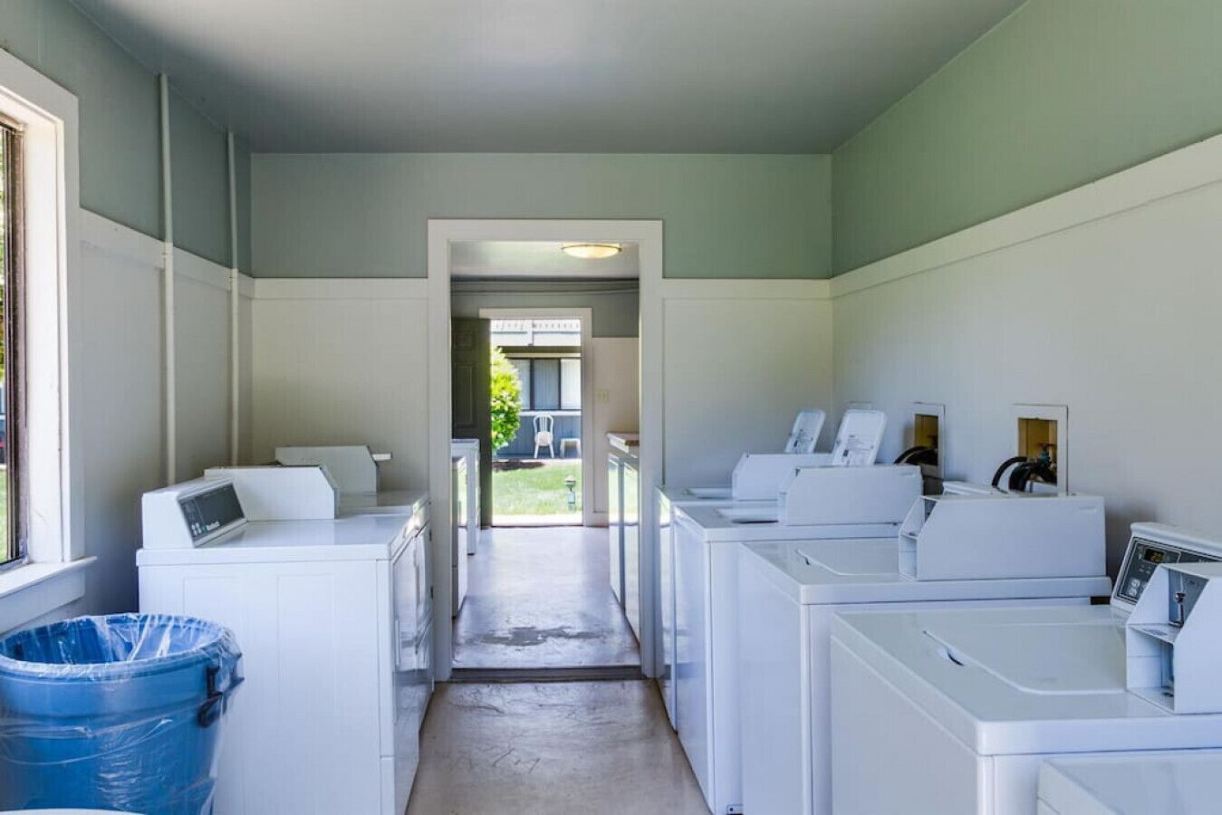 A laundry room with multiple washing machines and dryers, a blue trash bin, and a concrete floor.