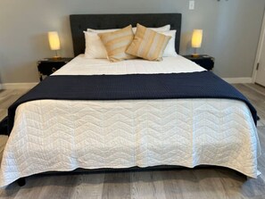 Un lit bien fait, recouvert d’une couette blanche et d’un plaid bleu foncé. Deux tables de chevet, chacune avec une lampe, sont placées de part et d’autre du lit.