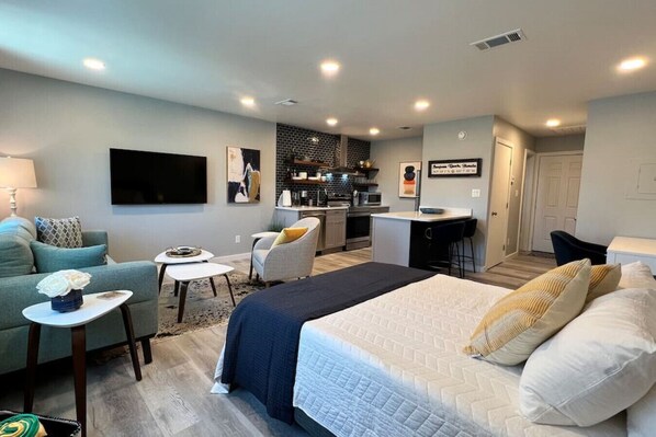 Une chambre d’hôtel moderne avec un grand lit, un coin salon comprenant un canapé et des fauteuils, une petite table à manger et, au fond, une kitchenette.