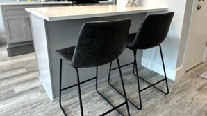 Ett modernt kök med en central köksö, två svarta barstolar och en vit bänkskiva.