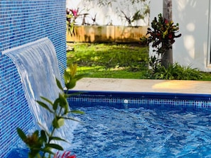 Une piscine agrémentée d’une cascade, entourée d’un jardin et d’une clôture en bois.