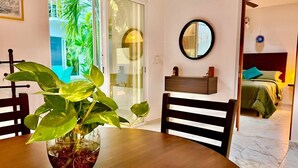Un área de comedor con una mesa de madera, sillas y una planta en maceta. Se ve una puerta corrediza de vidrio que da a un balcón con vegetación. Al fondo, una mesita de noche con una lámpara y un espejo.