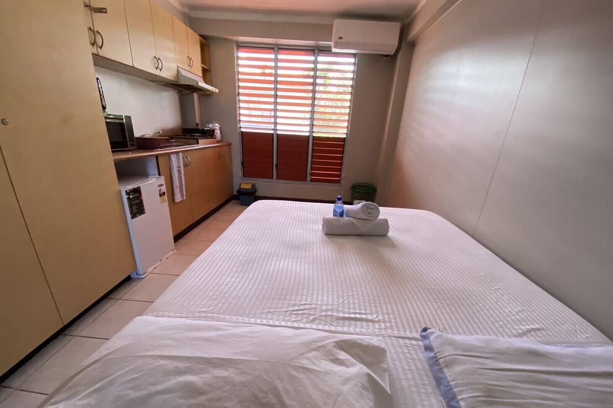 A compact hotel room with a bed, kitchenette, and a window with blinds.