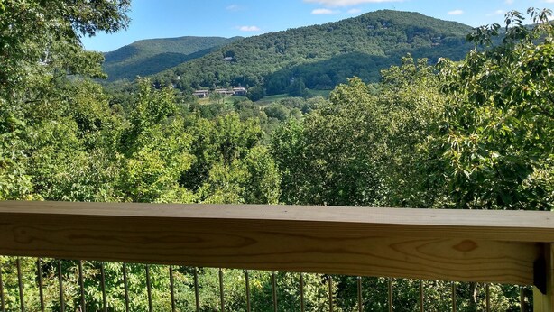 Uitzicht vanaf een balkon met houten hekwerk, over een weelderig groen landschap met een heuvel en verspreide gebouwen.