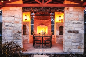 An outdoor stone fireplace with a table and chairs, surrounded by stone walls and decorative lanterns. - Blue Haus | 3/2 | Hot Tub, Outdoor Entertainment Area (Fredericksburg)