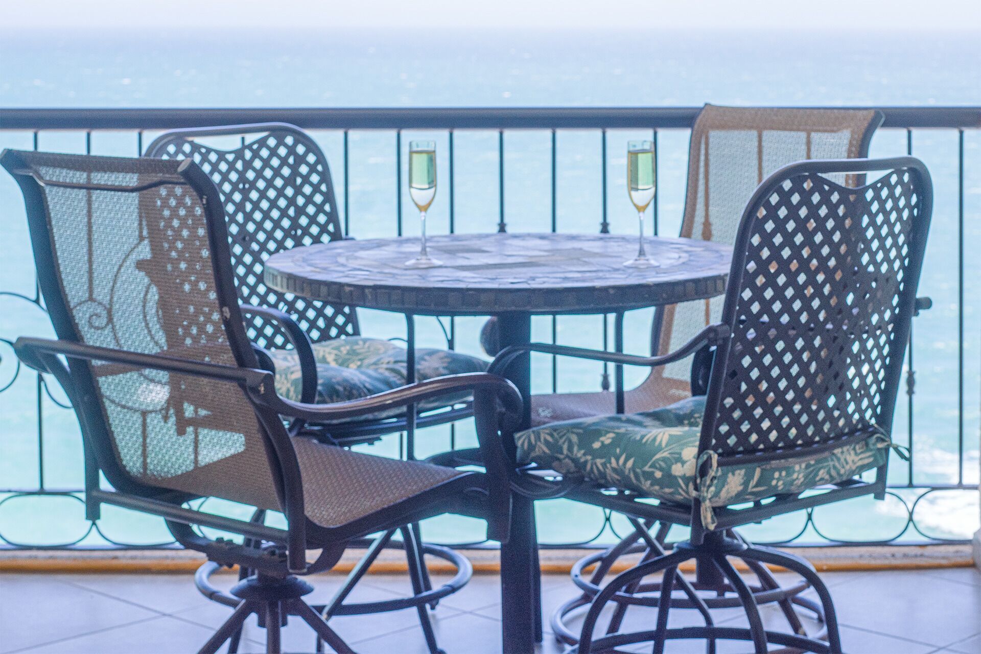 A round table with four chairs, each with a mesh backrest and cushioned seat, on a balcony with a view of the sea.