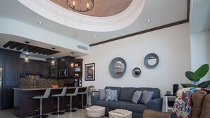 Eine moderne Küche mit dunklen Holzschränken, einer Granitabdeckung und einer Bar mit Hockern. Im Wohnbereich befindet sich eine graue Couch und ein Ottomanen, sowie eine große Pflanze in der Ecke.