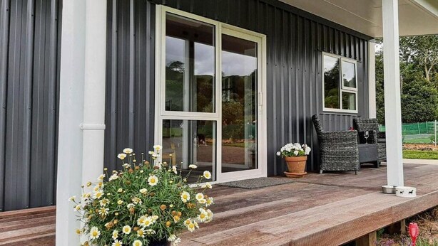 Une maison moderne avec une façade sombre, de grandes portes-fenêtres coulissantes, une terrasse en bois et du mobilier d’extérieur.