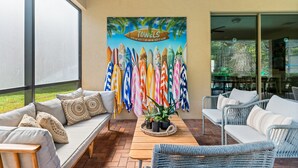 A sunlit patio with a sofa, wicker chairs, and a table with potted plants. A colorful wall art piece with surfboards is visible.