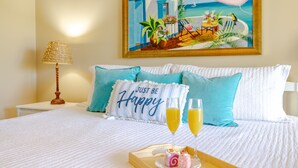 Cama bem arrumada com um travesseiro que diz 'Just Be Happy', dois copos de suco de laranja e uma bandeja com doces.